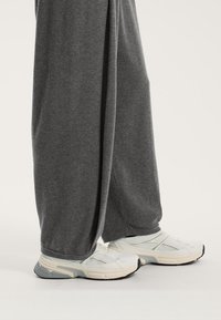 Sive trenirka hlače širokog kroja s glatkom teksturom, u kombinaciji s bijelim i sivim sportskim tenisicama koje imaju mrežaste detalje i gumene potplate.