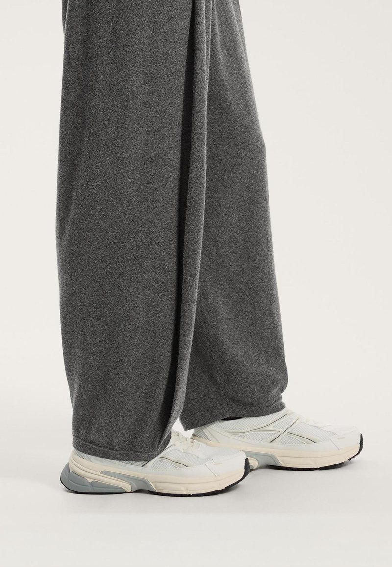 Sive trenirka hlače širokog kroja s glatkom teksturom, u kombinaciji s bijelim i sivim sportskim tenisicama koje imaju mrežaste detalje i gumene potplate.