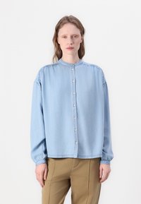 Lys blå denimskjorte med stående krage, lange ermer og knappestilling foran. Har detaljer med rynker på skuldrene og mansjettene.