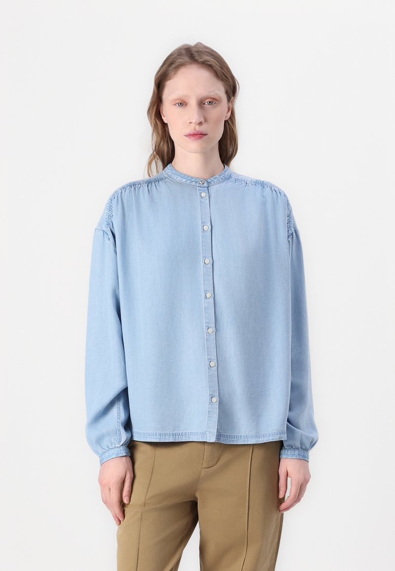 Lys blå denimskjorte med stående krage, lange ermer og knappestilling foran. Har detaljer med rynker på skuldrene og mansjettene.