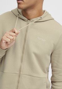 Bistezna zip-up dukserica napravljena od mekane tkanine, s kapuljačom na vezanje i malim otisnutim logom na prsima. Glatka tekstura, funkcionalan dizajn.