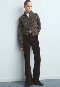 Bruine gebreide vest met ritssluiting en hoge kraag, gecombineerd met een donkerbruine, uitlopende broek met hoge taille. Model staat tegen een lichte muur.