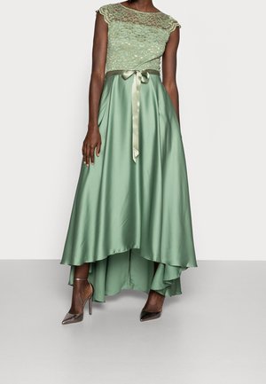 Woman wearing a green lace cap-sleeve top with a satin high-low skirt tied with a ribbon, and metallic pointed high heels.