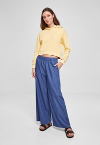 Felpa gialla corta con tasca a marsupio, abbinata a pantaloni in denim blu a gamba larga con cintura elastica e tasche laterali. Sandali neri.
