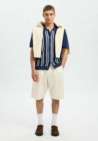 Kratka srajca s kratkimi rokavi in gumbi z mornarsko modrimi in belimi navpičnimi črtnimi vzorci, v kombinaciji s smetanastimi kratkimi hlačami. Smetanasta pulover se spusti preko ramen.