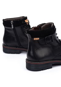 Botas de tobillo de cuero negro con un cuello de ante, cremallera lateral y detalle de hebilla, que cuentan con una suela texturizada y costuras blancas en contraste.