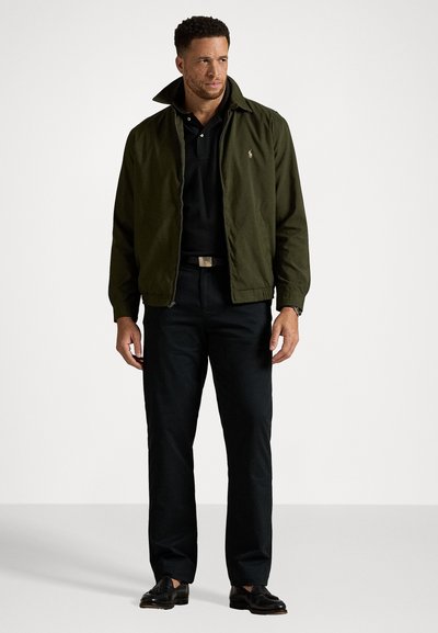 Veste aviateur verte olive avec fermeture éclair, portée sur un polo noir. Associée à un pantalon foncé et des chaussures en cuir noires.