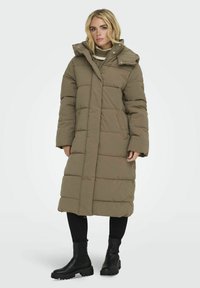 Manteau matelassé kaki avec un col montant, des boutons-pressions et un piquage horizontal. Porté sur un pull en tricot et un pantalon noir, associé à des bottes noires.