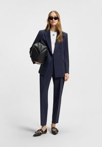 Navy tailored suit with double-breasted blazer, white top featuring black text, black handbag, and pointed-toe black shoes with straps.