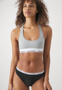 Soutien desportivo cinzento em algodão com uma faixa branca larga com a inscrição "Calvin Klein". Fio dental preto em renda transparente com detalhes semelhantes em faixa branca.