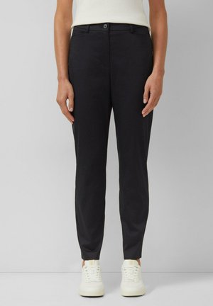 Kobieta ubrana w czarne dopasowane spodnie, białe tenisówki i biały bezrękawnik, stojąca na tle jednolitego jasnego tła.