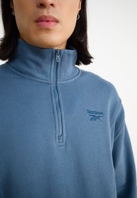 Pullover in pile con zip di colore azzurro chiaro, dotato di un colletto alto e con il logo Reebok ricamato in una tonalità di blu più scura. Texture morbida.