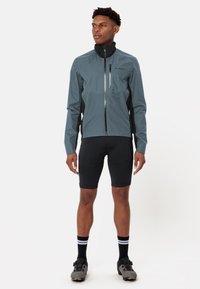 Veste de cyclisme imperméable grise avec un col haut, des panneaux latéraux noirs et une fermeture éclair à l'avant. Portée par-dessus un short noir et des chaussures de cyclisme.