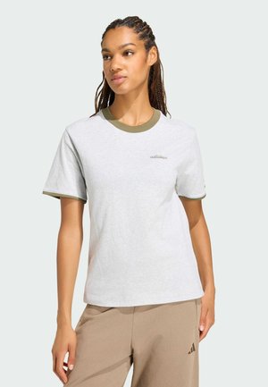Kvinde med flettet hår iført en hvid Adidas T-shirt med grøn kant og beige Adidas bukser, stående mod en ensfarvet baggrund.
