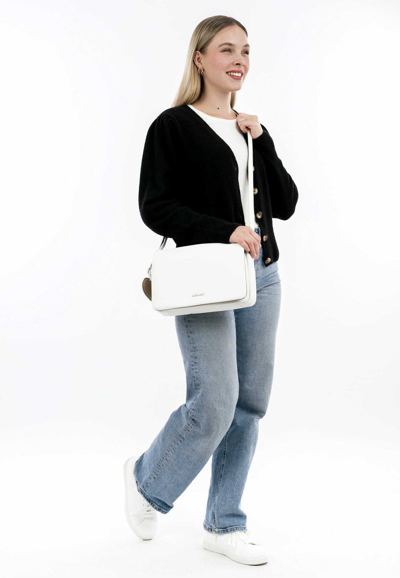 Hvid håndtaske med glat tekstur, sølvhardware og et aftageligt brunt accent. Båret af en person i en sort cardigan og blå jeans.