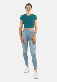 Türkisfarbener, kurzer T-Shirt mit kurzen Ärmeln, kombiniert mit hellblauen, hochgeschnittenen Jeans und weißen Sneakers, vor einem einfarbigen Hintergrund stehend.