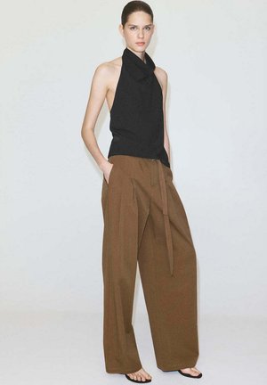 Femme aux cheveux attachés portant un haut noir sans manches à col halter et un pantalon large marron avec ceinture, debout les mains dans les poches.