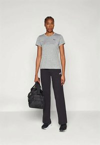 Žena stojí, oblečená v šedej tričku Puma, čiernych nohaviciach Puma a čiernych teniskách, v pravej ruke drží čiernu športovú tašku na pozadí s jednofarebným pozadím.