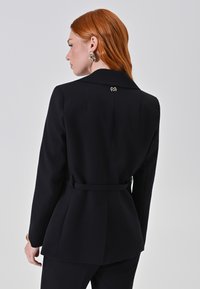 Blazer nero su misura con una silhouette strutturata, dettaglio a cintura in vita e un logo accentato in oro sul colletto. Tessuto dalla texture liscia.