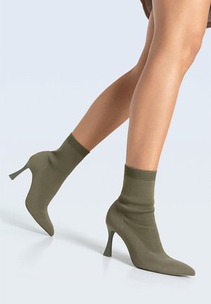 Jambes portant des bottines à talons hauts pointues de style chaussette de couleur vert olive sur un fond uni.