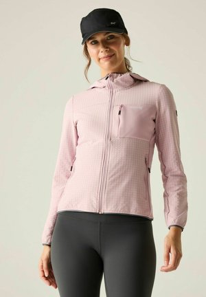 Mujer con una chaqueta rosa claro con cremallera, con bolsillos en el pecho y los laterales, y una gorra negra, de pie frente a un fondo liso y claro.