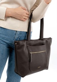 Brūna ādas tote soma ar strukturētu virsmu, kurai ir priekšējā rāvējslēdzēja kabata un izturīgas rokturi, attēlota uz gaišas apģērba un džinsu fona.