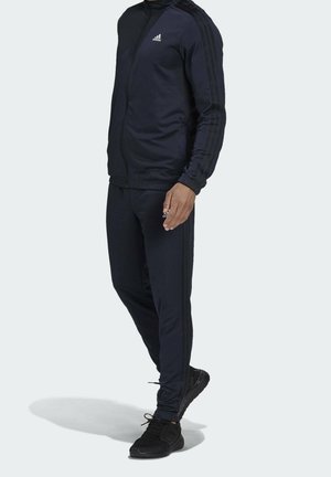 Man wearing navy blue Adidas track jacket and matching pants with black athletic shoes, standing against plain background.