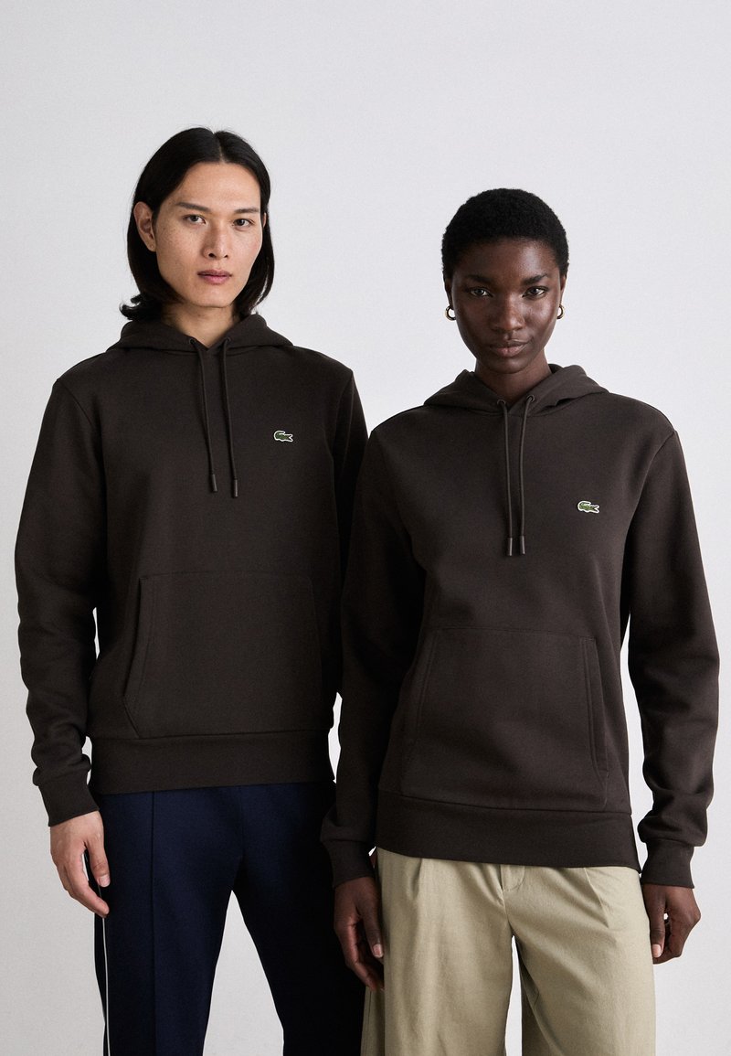 Sudaderas de algodón marrón con bolsillos delanteros y puños acanalados. Presenta un pequeño logo verde en el pecho. Los modelos llevan pantalones en navy y caqui claro.