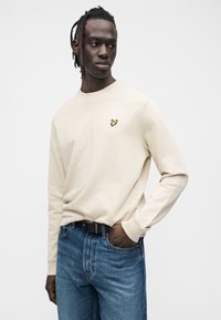 Béžová mikina s okrúhlym výstrihom s vyšívaným žltým logom na ľavej strane hrude, nosená s modrými džínsami a čiernym opaskom. Hladká textúra.