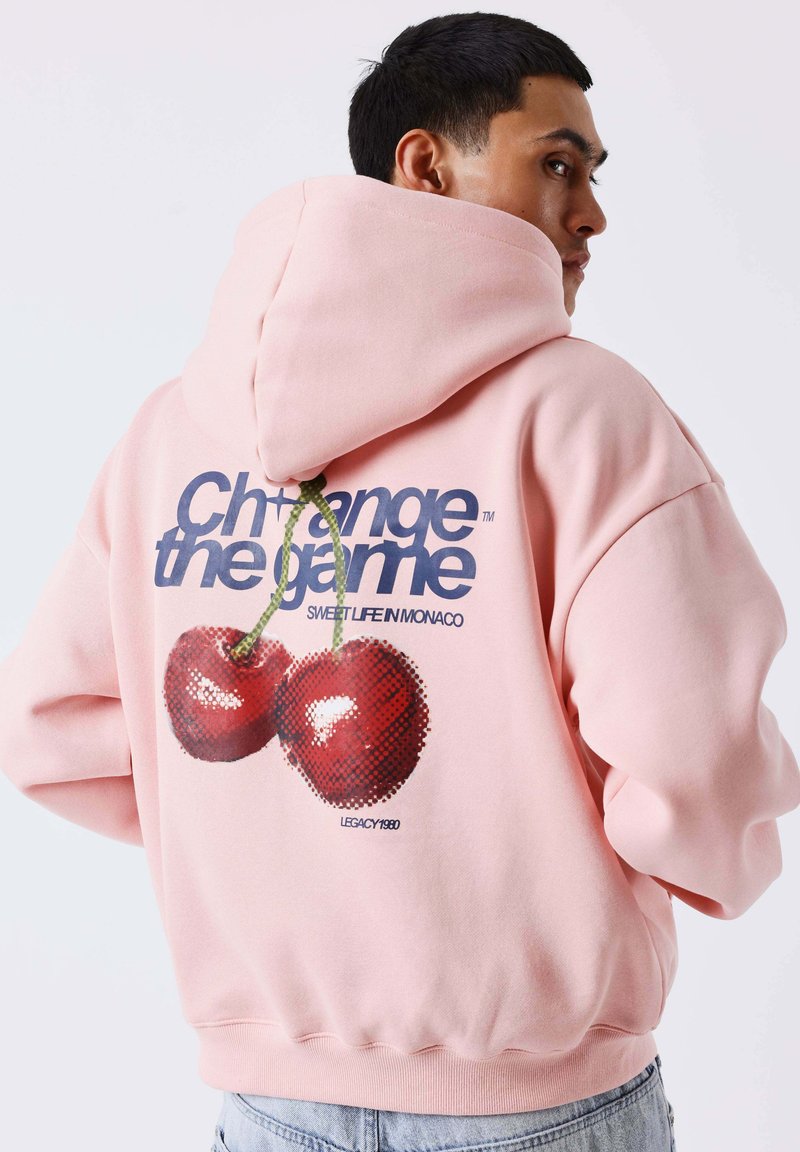 Homme portant un sweat à capuche rose avec un motif cerise et le texte « Change the game » dans le dos, regardant sur le côté, fond clair.
