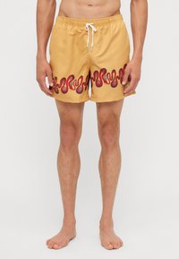Gelbe Badehose mit einem wellenförmigen roten und schwarzen Muster am Saum. Verfügt über einen elastischen Bund und eine Kordel zum Verstellen.