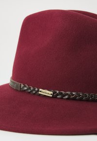 Burgundy színű filc fedora kalap széles karimával és fonott sötétbarna bőrszalaggal, amelyen egy kis arany Barbour logóplakett található.