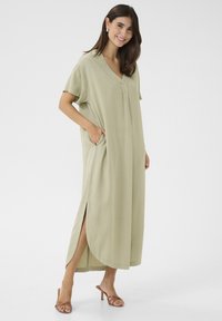 Térd hosszúságú, világos zöld ruha puha anyagból, V-nyakkivágással, rövid ujjal, oldalsó zsebekkel és oldalsó hasítékokkal a könnyű mozgás érdekében.