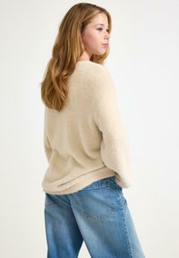 Beige pörröinen neule, jossa on pitkä hihainen malli, rento istuvuus ja pyöreä helma, yhdistettynä vaaleansinisiin farkkuihin.