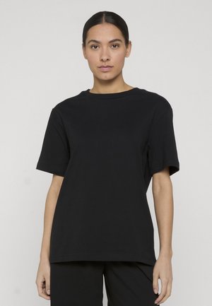Femme de face portant un T-shirt noir uni à manches courtes et un pantalon noir sur un fond gris clair.