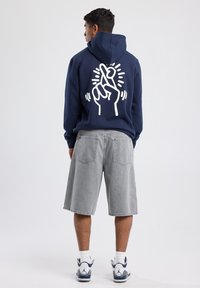 Sudadera con capucha azul marino con un diseño gráfico blanco de una mano haciendo un gesto en la parte de atrás, combinada con pantalones cortos grises y zapatillas deportivas.