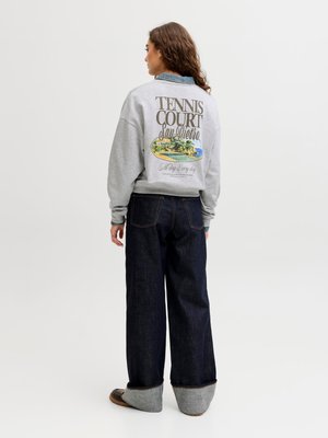 Sieviete ar muguru skatās, valkājot pelēku džemperi ar uzrakstu "Tennis Court San Pietro" un grafiku, saskaņojot to ar tumšām, plati piegrieztām atlocītām džinsa biksēm un brūnām kurpēm.