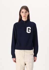 Marinblå stickad tröja med hög krage, dragkedja och vit "G"-logga på bröstet. Har ribbade ärmslut och nederkant.