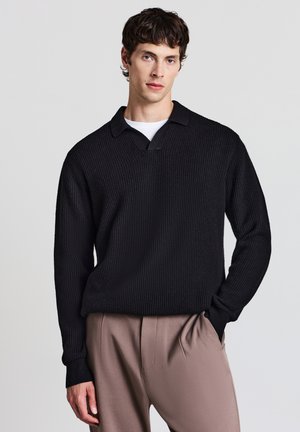 Pullover nero lavorato a maglia con scollo a V e colletto, caratterizzato da un tessuto testurizzato. Il modello indossa pantaloni taupe, con le mani in tasca.