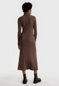 Rippstrickkleid in Dunkelbraun mit einem Stehkragen, langen Ärmeln und einer figurbetonten Silhouette, die bis zur Midi-Länge ausgestellt ist, kombiniert mit schwarzen Stiefeln.