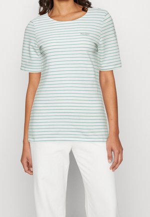 Kobieta w białej koszuli z cienkimi, poziomymi paskami w kolorze teal oraz logo "Marc O'Polo" na piersi, zestawiona z białymi spodniami.