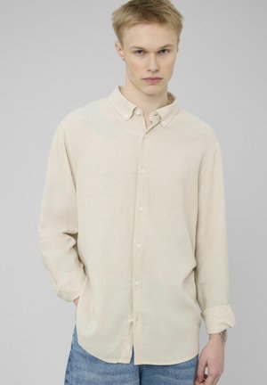 Jeune personne aux cheveux blonds courts portant une chemise beige à boutons aux manches retroussées et un jean bleu sur un fond uni.