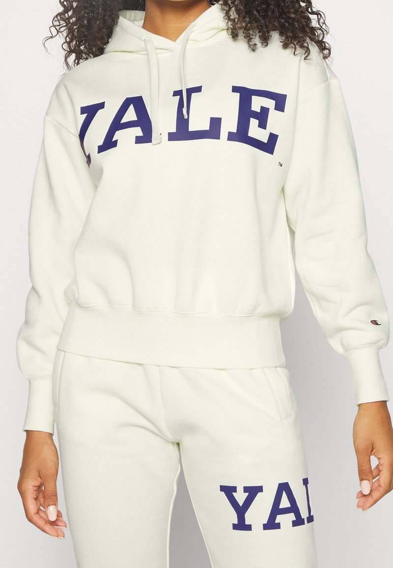 Ensemble sweat à capuche et pantalon de survêtement couleur crème avec grande inscription "YALE" en bleu marine. Tissu doux avec poignets et taille côtelés. Logo Champion sur la manche.