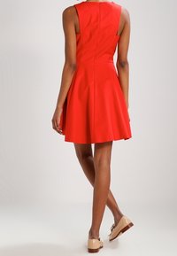 Vestido rojo sin mangas con falda acampanada, cierre de cremallera en la parte posterior y una textura suave. Combinado con zapatos planos de color claro.