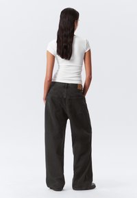 Los jeans negros de pierna ancha presentan una textura clásica de denim, dos bolsillos traseros y una discreta etiqueta con el logo en la cintura. Se combinan con una camisa blanca ajustada.