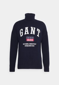 Dunkelblauer Strickpullover mit hohem Rollkragen, mit weißem Text "GANT" und einem Abzeichen mit Sternen und Streifen. Rippbündchen an den Ärmeln und am Saum.