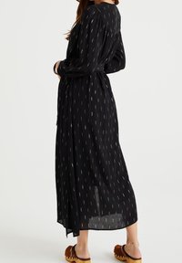 Černé dlouhé šaty s páskem v pase, mají skládací zadní stranu a diskrétní zlatý vzor po cel�ém povrchu. Přizpůsobeno s hnědými dřeváky.