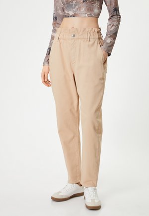 Pantalones de algodón beige con cintura alta, diseño plisado, bolsillos laterales y cierre de botón, combinados con zapatillas blancas y una blusa estampada.