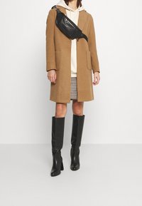 Abrigo de lana marrón, riñonera de cuero negro, sudadera blanca con capucha, minifalda de pata de gallo y botas de tacón altas negras, de pie sobre un fondo liso.