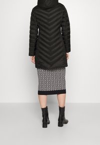 Schwarze Steppjacke mit Chevron-Quilt, Kapuze und langen Ärmeln. Getragen über einem knielangen Rock mit geometrischem Schwarz-Weiß-Muster.